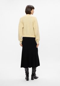 Pull en tricot jaune doux avec une coupe décontractée, associé à une jupe longue noire. Des bottes noires montantes viennent compléter le look, avec un talon épais.