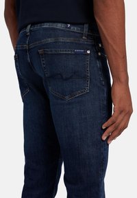 Jeans en denim bleu foncé avec une coupe slim, présentant des poches arrière avec des coutures visibles et un détail de logo subtil sur la ceinture.