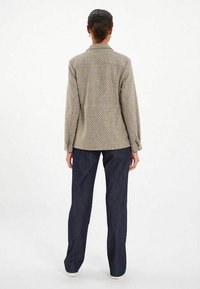 Chemise beige à motifs avec des manches longues, des poignets boutonnés et un col, associée à un jean droit bleu marine. Baskets blanches visibles.