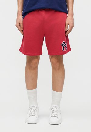Mand iført røde shorts med et navy-logo på venstre ben, hvide sokker, hvide sneakers og en navyblå skjorte, stående mod en ensfarvet baggrund.