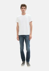 Fehér, rövid ujjú pamut póló, kék farmernadrággal és szürke tornacipővel, semleges háttér előtt állva.