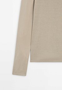 Manche et section du torse d'un pull en tricot beige à manches longues sur fond blanc, montrant la texture du tissu et une couture d'ourlet nette.