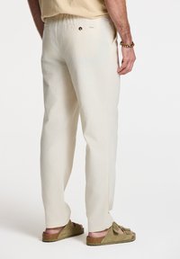 Pantalone od lana boje kreme sa opuštenim krojem, s jednim zadnjim džepom, u kombinaciji s maslinasto-zelenim kliznim sandalama. Glatka tekstura.
