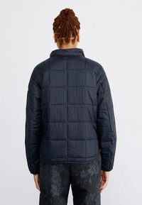 Personne aux cheveux tressés portant une veste matelassée noire et un pantalon sombre à motif floral, debout, dos au spectateur.
