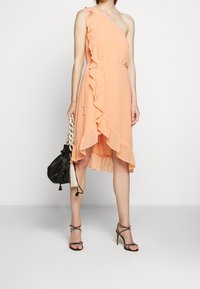 Robe pêche à épaule unique avec des volants sur le côté, ourlet asymétrique et tissu léger. Associée à des talons noirs à lanières et un sac à main.