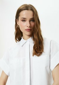 Camicia bianca a maniche corte con colletto button-down e un dettaglio della cucitura frontale. Tessuto di cotone liscio con una vestibilità rilassata. Nessun motivo o accento.