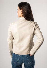 Beige leren jack met een getailleerde taille, ritsmanchetten en verticale naaddetails. Draag het over een blauwe denim jeans. Achteraanzicht.