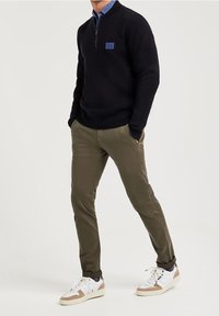 Pull marin à col zippé, pantalon kaki et baskets blanches avec des accents beige. Doté d'un patch logo contrastant sur le pull.