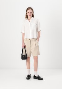 White short-sleeve linen shirt, beige tailored shorts, black bag with logo, black loafers, white socks, standing on a plain background.