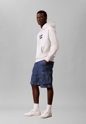 Ung mand stående, iført en hvid hættetrøje med tekst, blå cargobukser, hvide sokker og hvide sneakers mod en ensfarvet grå baggrund.