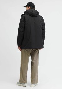 Sort isoleret jakke med hætte, der har en glat tekstur. Bæres over beige løstsiddende bukser og grå sneakers.