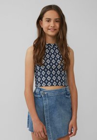 Navyblaues ärmelloses Cropped Top mit weißem geometrischem Muster; kombiniert mit einem hellblauen Denim-Wickelrock mit seitlichem Knopfverschluss und fransigem Saum.
