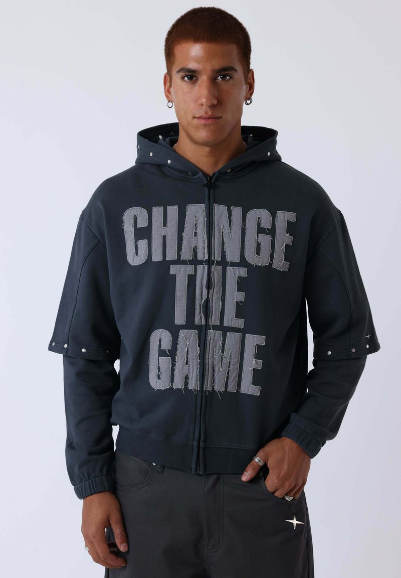 Jeune homme aux cheveux courts portant une veste à capuche foncée avec le texte "CHANGE THE GAME" et un pantalon noir orné d'un symbole d'étoile.