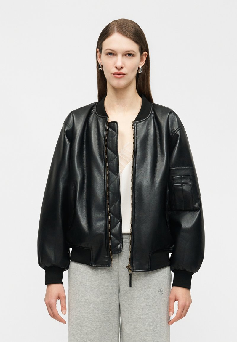 Femme portant une veste aviateur en cuir noir avec des poignets et un col côtelés, dézippée sur un haut blanc, associée à un pantalon gris clair.