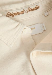 Κρεμ shirt με γιακά, φτιαγμένο από υφασμάτινη υφή, που διαθέτει χρυσή κεντημένη επιγραφή "Originals Studio" και λεπτομέρεια κουμπιού.
