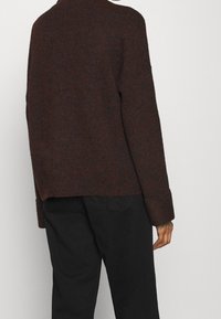 Ruskea neulepaita, jossa on korkea kaula-aukko, roikkuvat hartiat ja ribbireunat. Irtonainen muotoilu suoran helman ja teksturoidun kankaan kanssa.