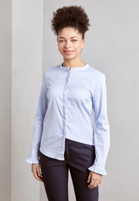 Camicia a maniche lunghe azzurro chiaro con apertura a bottoni, polsini arricciati e colletto arrotondato, abbinata a pantaloni scuri. Tessuto morbido, taglio su misura.