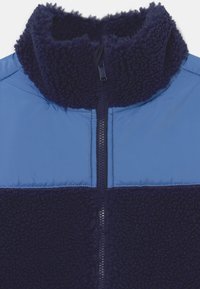 Dark blue fleece jacket with a high collar and light blue quilted fabric panel across the chest and shoulders, featuring a central zipper.