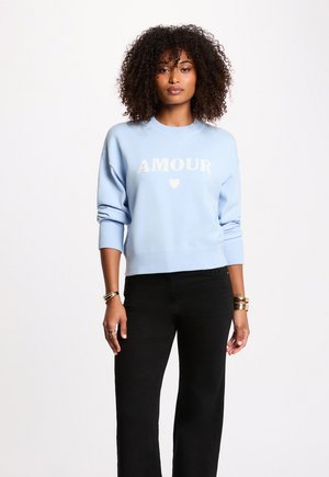 Kvinde med krøllet hår iført en lyseblå sweater med "AMOUR" og et hjerte-symbol, kombineret med sorte bukser og gyldne armbånd.
