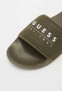 Olivgrüne Rutschsandalette mit einer strukturierten Gummisohle und einem atmungsaktiven Obermaterial aus Mesh, das das weiße "GUESS"-Logo und das Gründungsjahr zeigt.