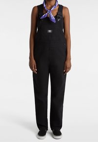 Schwarze Denim-Latzhose mit verstellbaren Trägern, einer Fronttasche und seitlichen Knöpfen. Kombiniert mit einem lila karierten Schal und schwarzen Turnschuhen.
