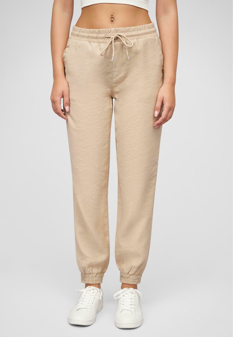 Beige jogger bikses, kas izgatavotas no mīkstas auduma, ar elastīgu jostasvietu un saiti, kā arī elastīgām aprocēm potītēs. Valkātas ar baltām sporta apaviem.