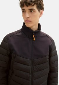 Czarna ocieplana kurtka z usztywnioną górną częścią, wysokim kołnierzem i pomarańczowym suwadłem. Posiada pikowaną strukturę oraz boczne kieszenie.