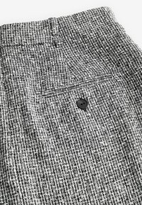 Texturovaná černobílá látka se jemným kostkovaným vzorem, obsahující černý knoflík a zadní kapsu na pase.