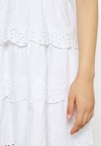 Pravá ruka spočívající vedle bílé šaty s vyšívanými květinovými výřezy na vrstvené textilii.