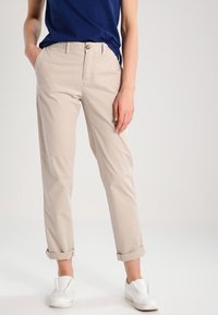 Person klädd i beiga upprullade byxor, en blå skjorta och vita slip-on skor, står med en hand i fickan mot en neutral bakgrund.