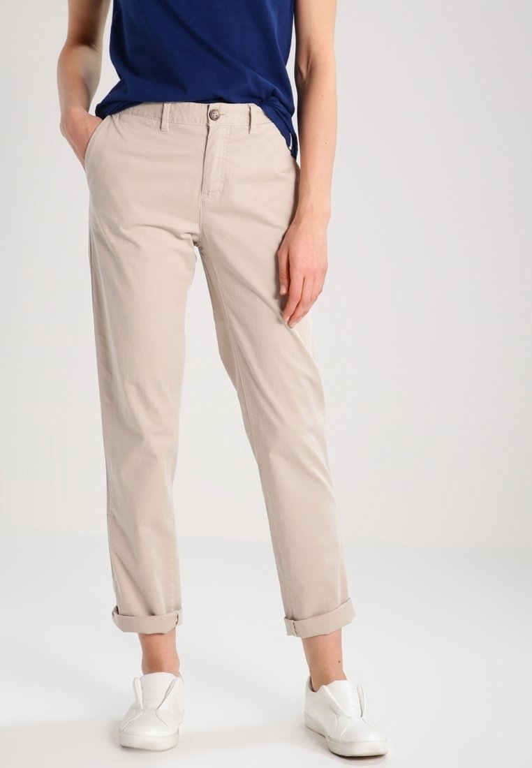 Person klädd i beiga upprullade byxor, en blå skjorta och vita slip-on skor, står med en hand i fickan mot en neutral bakgrund.