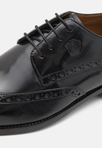 Scarpa nera in pelle con superficie liscia, decorazioni strutturate lungo il bordo e sei asole per i lacci. Presenta un dettaglio del logo in rilievo.