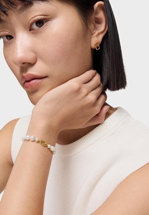 Femme aux cheveux noirs raides portant une boucle d'oreille en or en forme d'anneau et un bracelet de perles avec fermoir en or, vêtue d'un haut blanc côtelé sans manches.