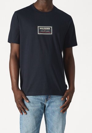 Mand iført navyblå T-shirt med rektangulært logo-patch med teksten "Hilfiger American Classic Est. 1985 New York" og lyseblå jeans.