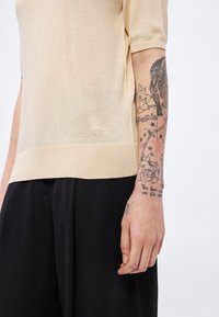 Top bej tricotat cu mâneci scurte, tiv și placket cu model tip dungi. Include un logo brodat subtil și brațul tatuat parțial vizibil al unui model.