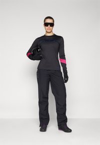 Svart langermet skjorte med blå og rosa detaljer, kombinert med svarte bukser og hansker. Modellen har solbriller på seg og holder en hjelm.