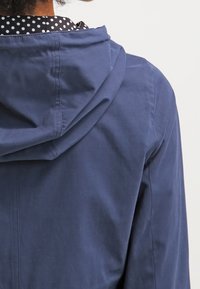 Marinblå hoodie-jacka med en slät yta, med en detaljerad prickig foder som syns vid kragen och ryggen.