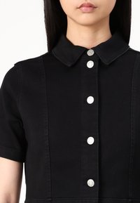 Vestido de ganga preto com colarinho, mangas curtas, cinco botões de pressão prateados na frente, apresentando detalhes de costura vertical e uma cintura ajustada.