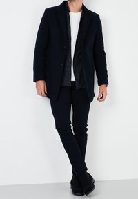 Tmavě modrý kabát z vlněné směsi, úzké tmavé kalhoty, bílé tričko a černé kotníkové boty. Jednoduchý design s knoflíkovými detaily a límcem.