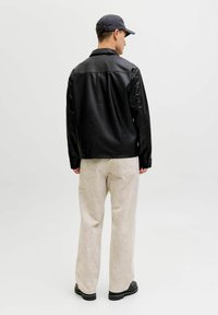 Schwarze Lederjacke mit glatter Textur, Knopfverschluss und langen Ärmeln. Kombiniert mit hellbeigen, weit geschnittenen Hosen und schwarzem Schuhwerk.