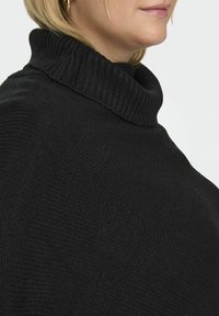 Poncho en tricot noir avec un col montant côtelé, tissu texturé et design ample, mettant l'accent sur la chaleur et le confort.