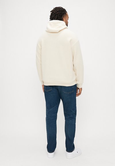 Κρέμ φλις φούτερ με υψηλό λαιμό, συνδυασμένο με σκούρο μπλε τζιν παντελόνια και λευκά αθλητικά παπούτσια, εμφανίζεται από πίσω.