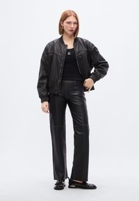 Μαύρο δερμάτινο μπουφάν bomber με πλεκτά τελειώματα, συνδυασμένο με εφαρμοστά μαύρα δερμάτινα παντελόνια. Το μοντέλο φοράει μαύρο αμάνικο τοπ με λογότυπο και μαύρα μοκασίνια.