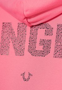 Růžová mikina s velkými, texturovanými černými rhinestony, které po celé přední části tvoří slovo "DANGER", s malým logem pod textem.
