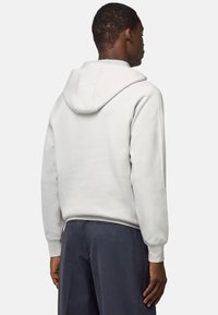 Felpa grigia chiara con cucitura centrale sulla schiena e un grande cappuccio tondo. Tessuto morbido con polsini a costine, indossata con pantaloni blu navy.