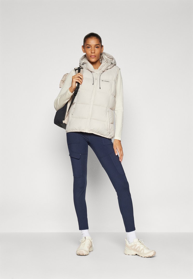 Puffer vest in lichtbeige met capuchon, volledige rits aan de voorkant en logo-detail. Gepaard met marineblauwe cargo leggings en lichte sneakers.