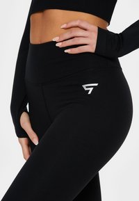 Black high-waisted leggings made from stretchy fabric, featuring a slim fit and a silver brand logo on the left thigh. Seam detailing visible.