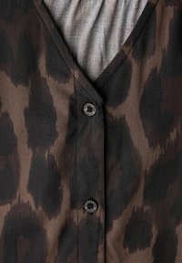 Blusa marrón con estampado de leopardo que presenta un escote en V, tela suave y un botón negro con detalle de logo en la tapeta. La textura parece suave.