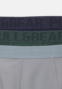 Tri páry pánskeho spodného prádla s kontrastnými pásikmi: tmavomodré, zelené a svetlosivé, každé s embosovaným nápisom "PULL&BEAR" v zodpovedajúcich farbách.