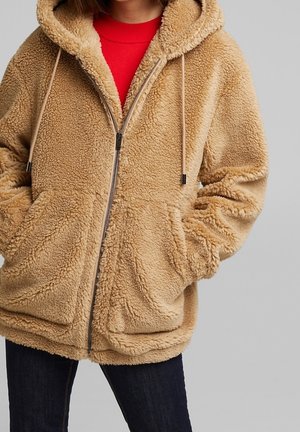 Persoon draagt een beige zip-up fleece hoodie met voorzakken en een rood shirt eronder, zichtbaar van borst tot heupen.